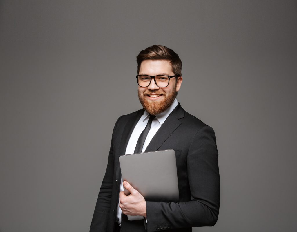Portrait of a happy young businessman dressed in suit holding laptop computer and looking at camera isolated over gray background
